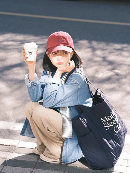 Original photo: a young woman in a red cap and denim jacket squatting on a sidewalk with a coffee cup and a navy tote bag